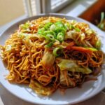 A plate of savory, saucy fried noodles mixed with cabbage and carrots, topped with fresh sliced green onions.