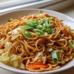 A close-up of a white bowl filled with saucy, brown fried noodles mixed with cabbage and carrots, topped with green onions.