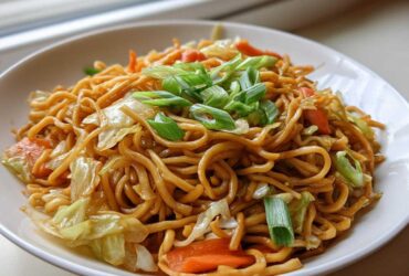 A close-up of a white bowl filled with saucy, brown fried noodles mixed with cabbage and carrots, topped with green onions.