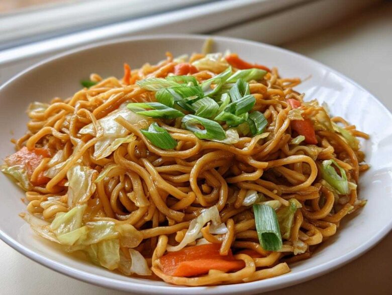 A close-up of a white bowl filled with saucy, brown fried noodles mixed with cabbage and carrots, topped with green onions.