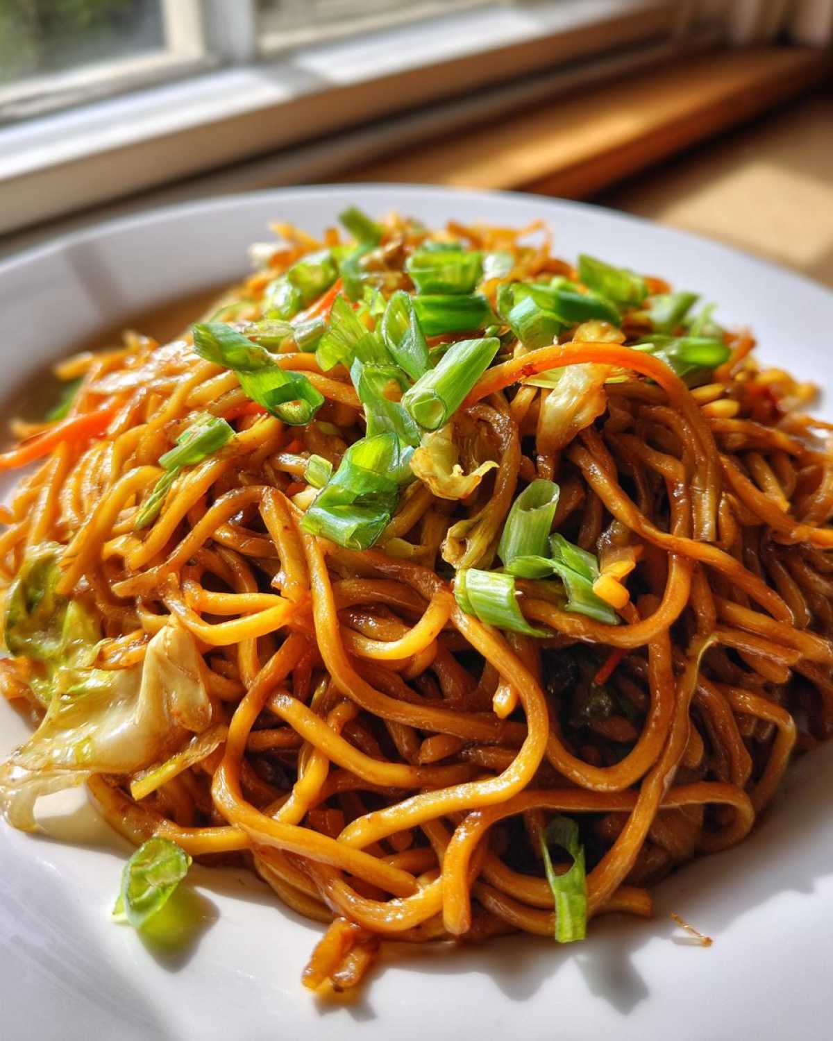 A close-up, brightly lit serving of glossy, brown fried noodles topped generously with fresh green chopped scallions.