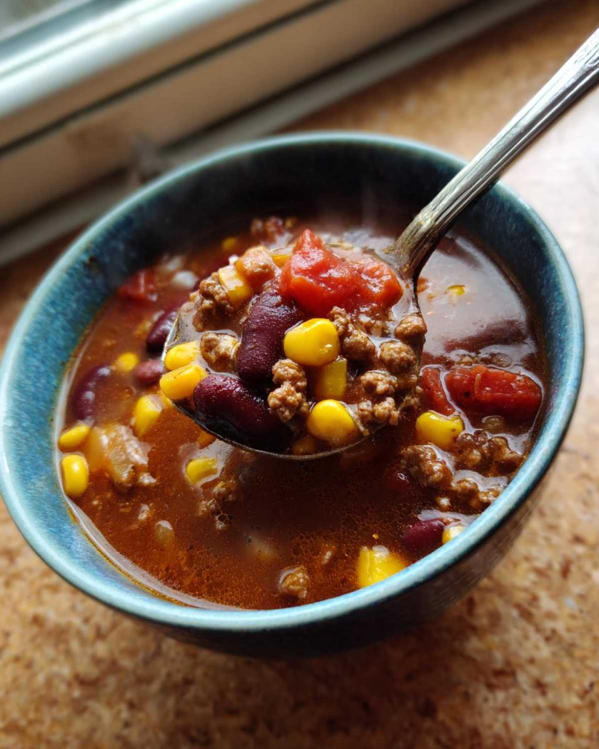 A spoonful of hot crockpot cowboy soup, featuring ground beef, kidney beans, corn, and tomatoes, being lifted from a blue bowl.