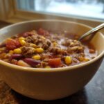 A steaming bowl of hearty crockpot cowboy soup featuring ground meat, kidney beans, corn, and tomatoes, set near a window.