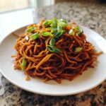 A plate of sticky garlic noodles coated in a dark sauce and garnished with fresh chopped green onions.