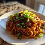 A close-up of a portion of sticky garlic noodles coated in a dark sauce and generously topped with bright green chopped scallions.