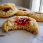 Close-up of a strawberry cheesecake cookies with a bite taken out, showing the soft interior and bright red jam filling.