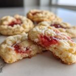 A close-up view of soft strawberry cheesecake cookies, with one cookie broken in half showing the creamy texture and bright red strawberry filling.