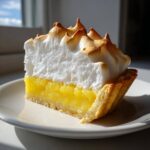 A tall slice of lemon meringue pie showing bright yellow filling and perfectly peaked, lightly browned meringue topping.