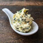 A dollop of whipped garlic butter mixed with green herbs served in a small white tasting spoon.