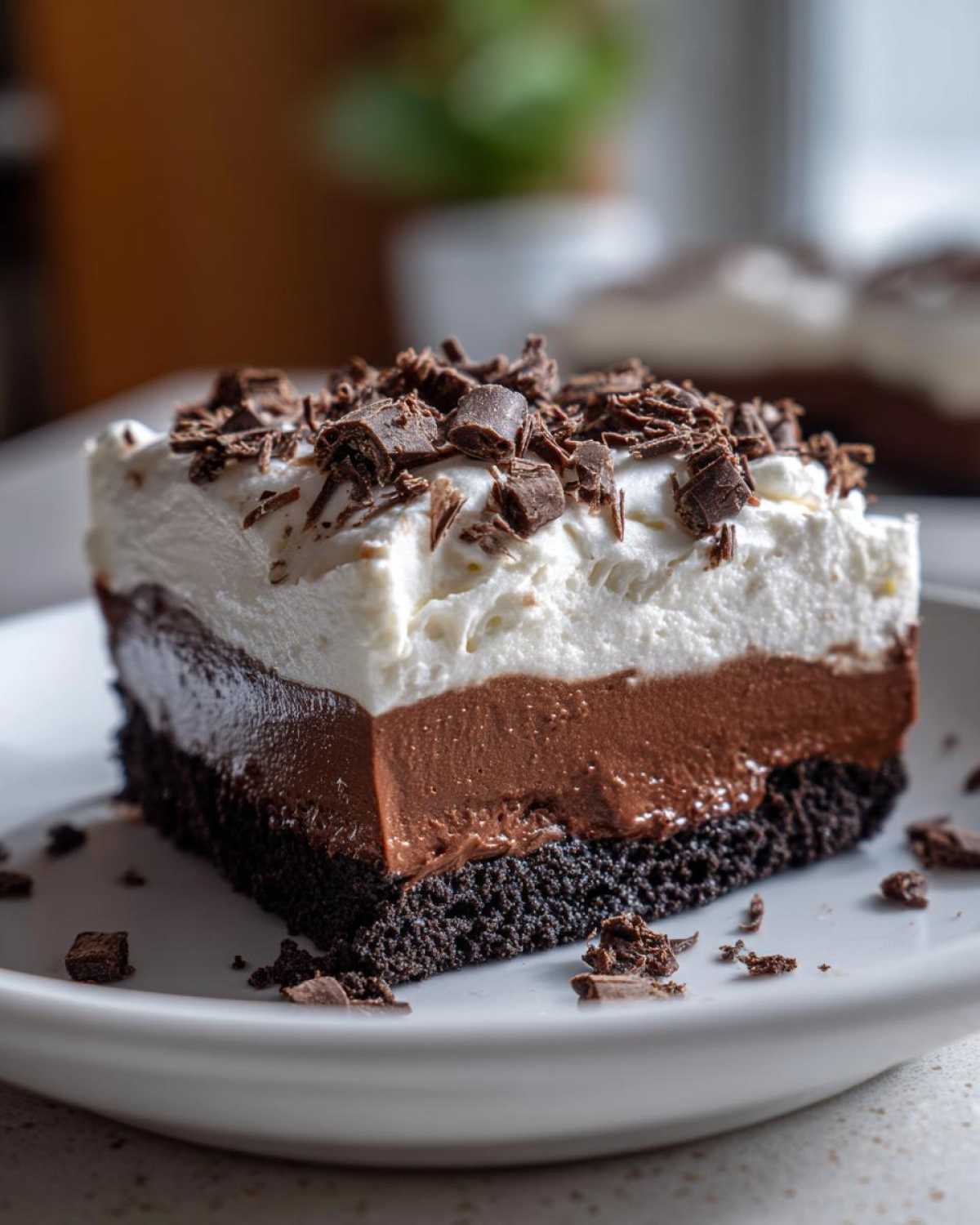 A square slice of layered chocolate mud pie featuring a dark crust, chocolate pudding, whipped topping, and chocolate shavings.