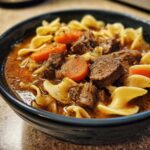 Close-up of a dark bowl filled with rich beef noodle soup, featuring tender beef chunks, wide egg noodles, and sliced carrots.