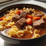 Close-up of a rich beef noodle soup featuring tender beef chunks, egg noodles, and bright orange carrots.