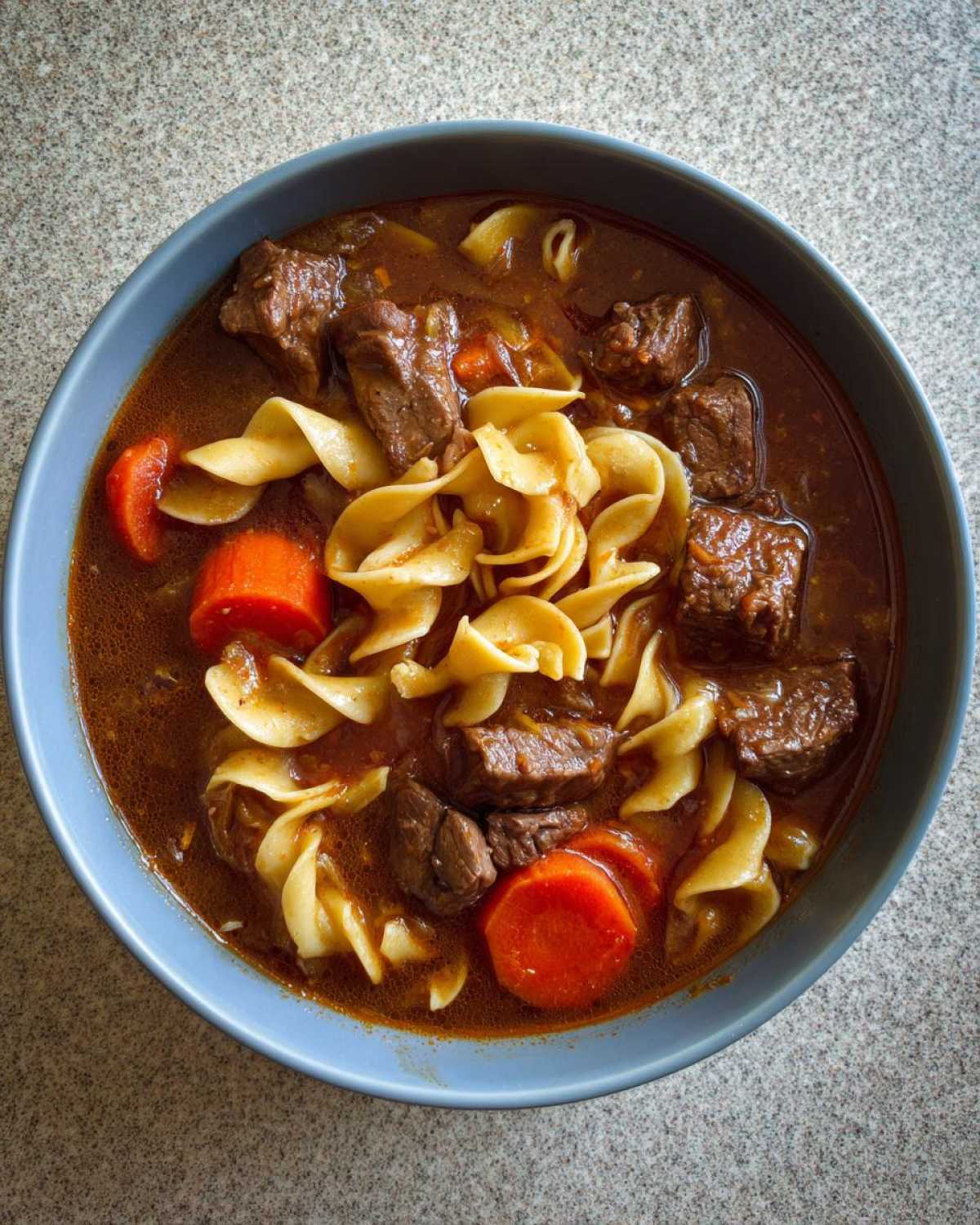 Overhead view of a bowl of rich beef noodle soup with chunks of beef, carrots, and egg noodles.