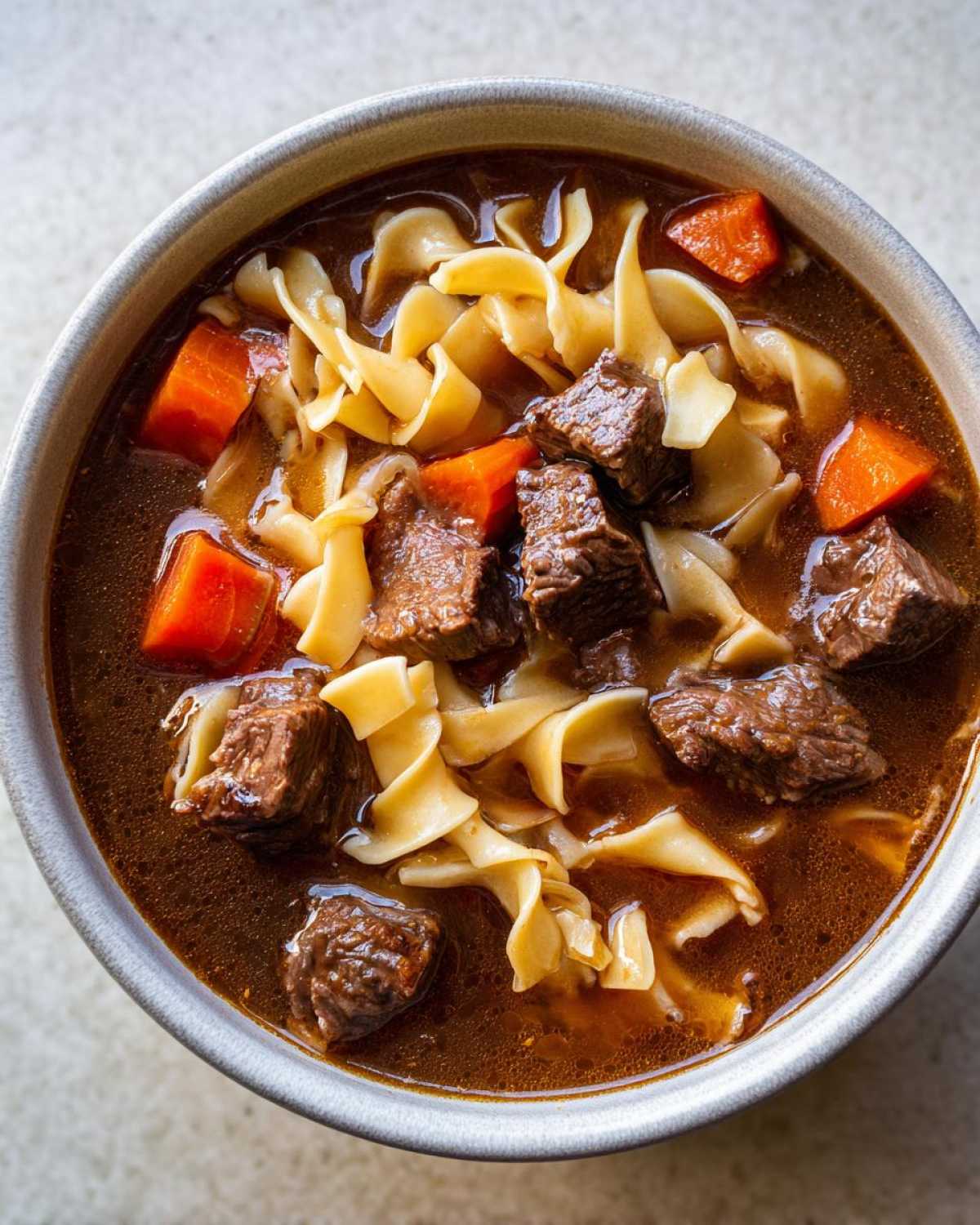 Overhead close-up of rich beef noodle soup with chunks of beef, egg noodles, and carrots in a grey bowl.