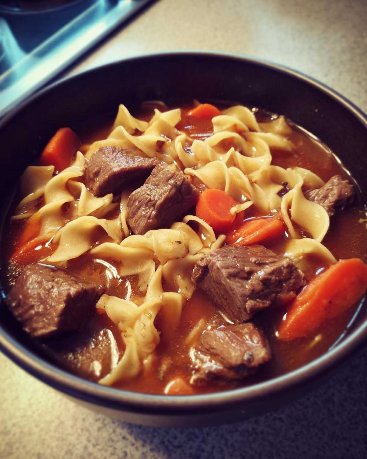 Close-up of a dark bowl filled with rich beef noodle soup, featuring chunks of beef, egg noodles, and sliced carrots.