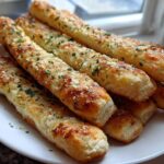 A close-up of several golden, cheesy breadsticks topped with garlic and parsley served on a white plate.