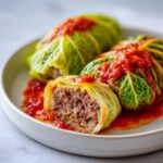 Close-up of three cabbage rolls served on a white plate, one cut open to show the savory meat filling, topped with tomato sauce.