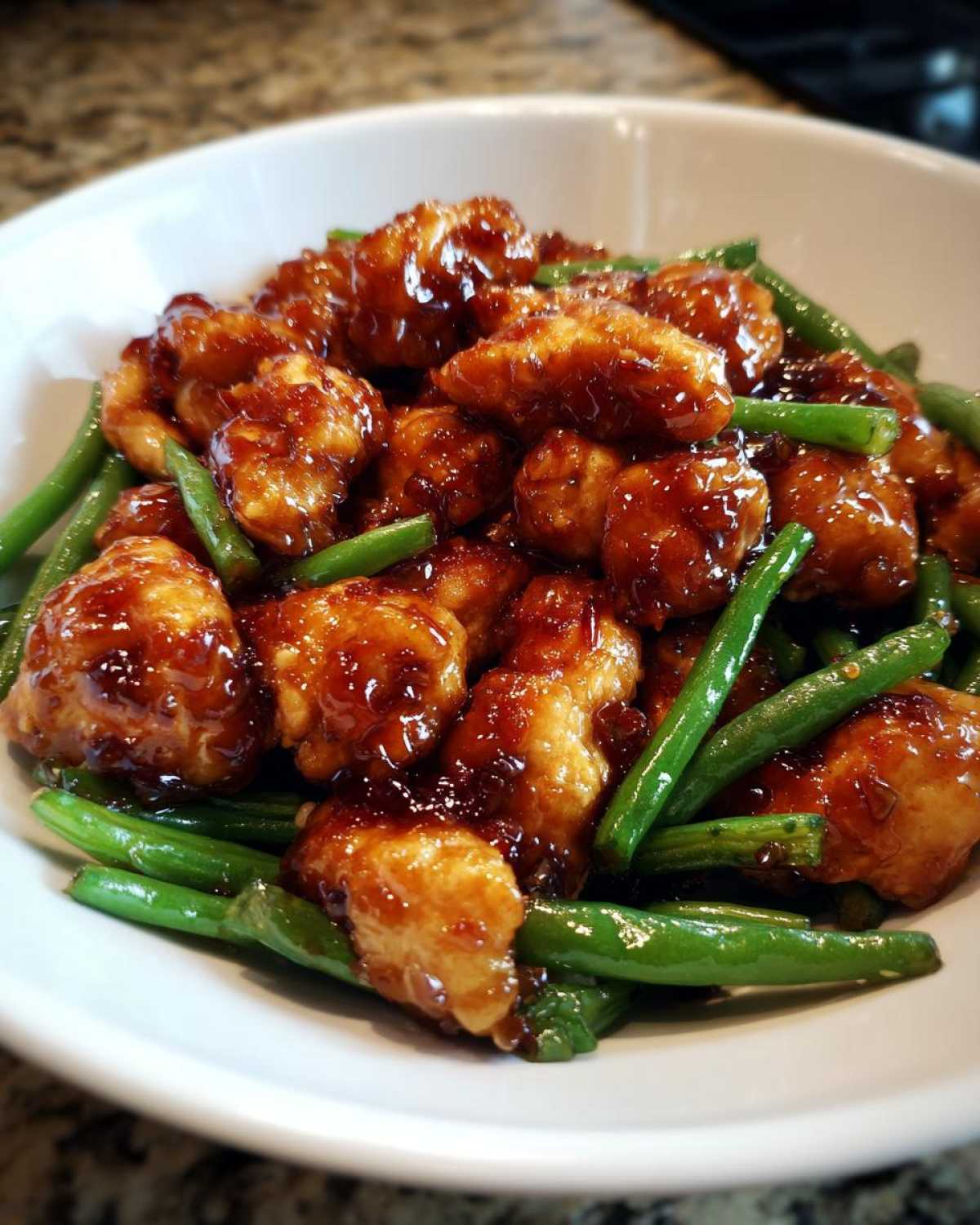 Close-up of crispy chicken pieces coated in a dark, glossy sauce mixed with bright green beans, served in a white bowl.
