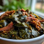 A close-up of tender, slow-cooked collard greens topped with shredded smoked meat in a white bowl.
