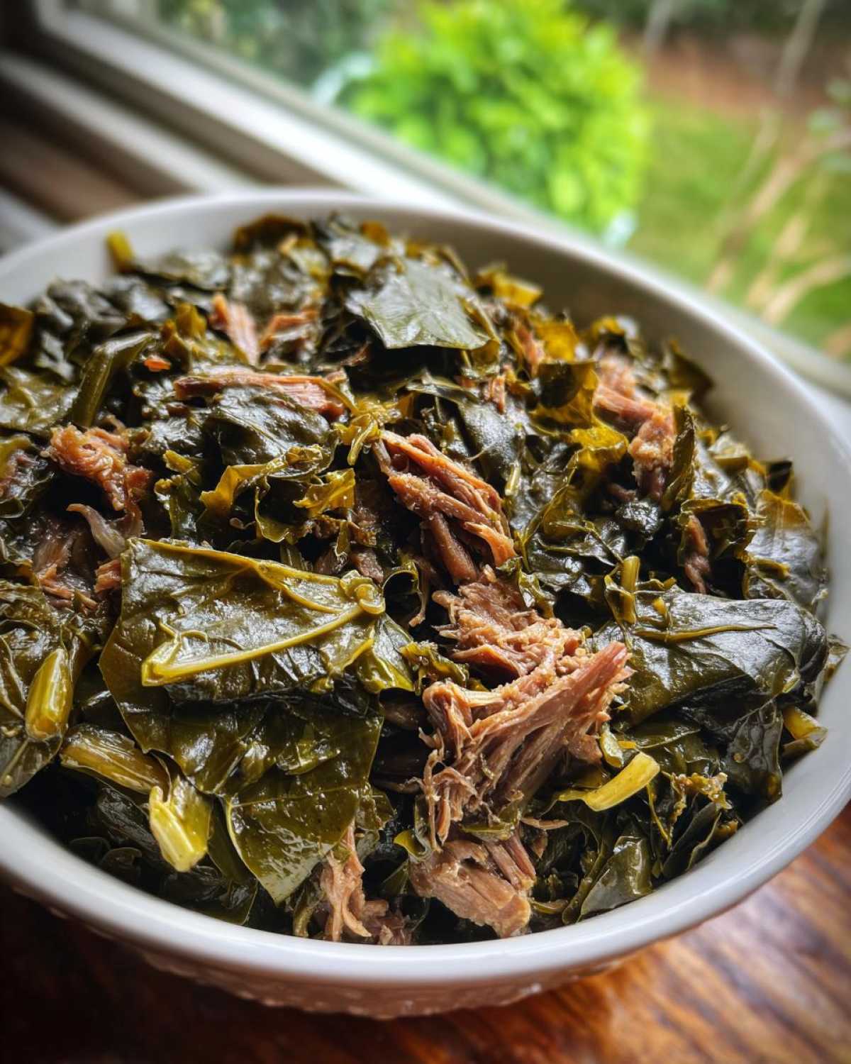 Close-up of a white bowl filled with tender, dark green collard greens mixed with shredded smoked meat.