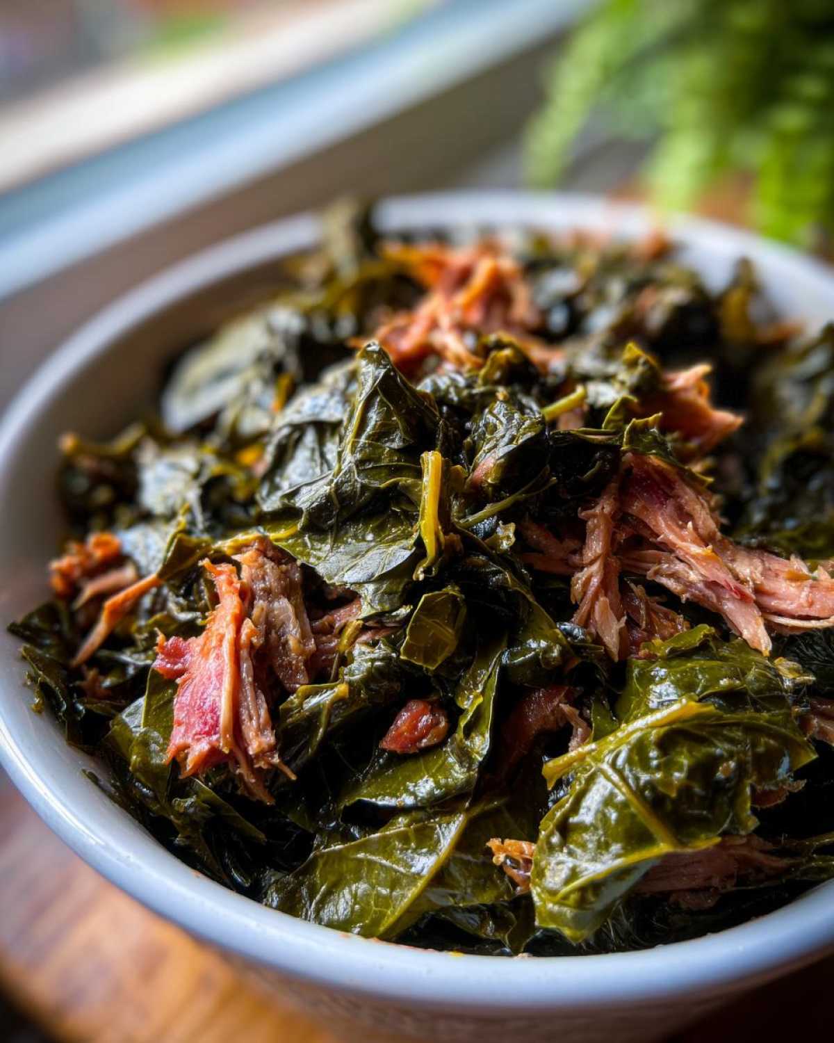 Close-up of tender, dark green collard greens mixed with shredded smoked meat in a white bowl.