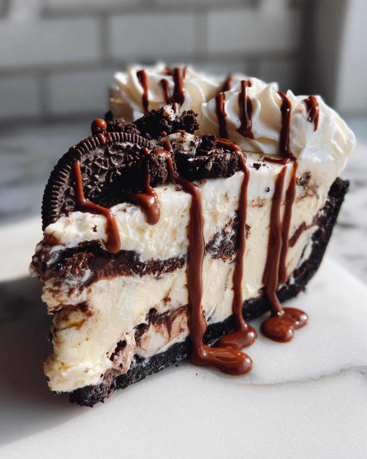 A decadent slice of ice cream pie with layers of cream, cookie crust, whipped topping, and chocolate syrup drizzle.