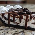 Close-up of a decadent slice of ice cream pie with an Oreo crust, vanilla filling, chocolate fudge drizzle, and whipped cream.