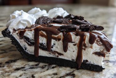 Close-up of a decadent slice of ice cream pie with an Oreo crust, vanilla filling, chocolate fudge drizzle, and whipped cream.