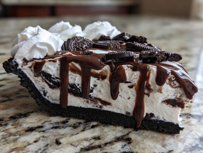 Close-up of a decadent slice of ice cream pie with an Oreo crust, vanilla filling, chocolate fudge drizzle, and whipped cream.