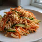 A close-up of a vibrant kani salad featuring shredded crab, julienned carrots, and sliced cucumbers, served on a white plate.