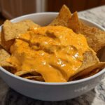 A white bowl filled with tortilla chips topped generously with creamy, orange nacho cheese dip.