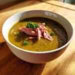 A close-up of a bowl of thick, green pea soup topped with shredded ham and fresh parsley.