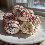 A pile of white chocolate coated sugar cookie truffles covered in rainbow sprinkles, one is broken open.