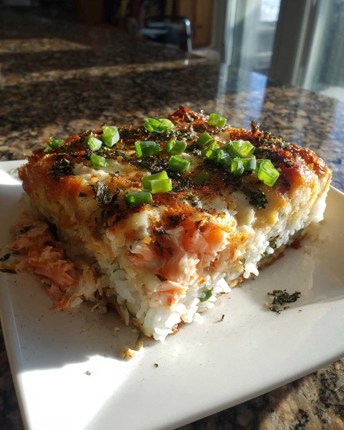 A square slice of baked sushi with layers of rice, salmon, and creamy topping, garnished with green onions.
