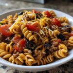 A close-up view of a white bowl filled with amazing taco pasta salad featuring rotini pasta, ground meat, tomatoes, olives, and cheese.