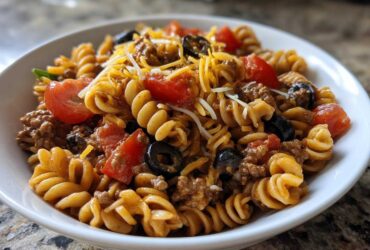A close-up view of a white bowl filled with amazing taco pasta salad featuring rotini pasta, ground meat, tomatoes, olives, and cheese.