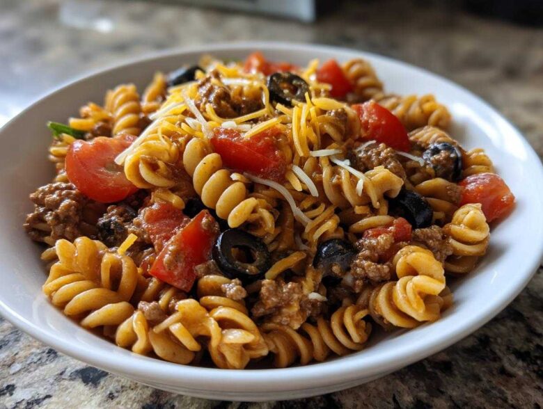 A close-up view of a white bowl filled with amazing taco pasta salad featuring rotini pasta, ground meat, tomatoes, olives, and cheese.