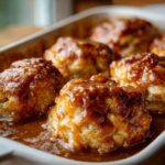 Close-up of several baked apple dumpling portions soaking in a rich, glossy caramel sauce in a white baking dish.