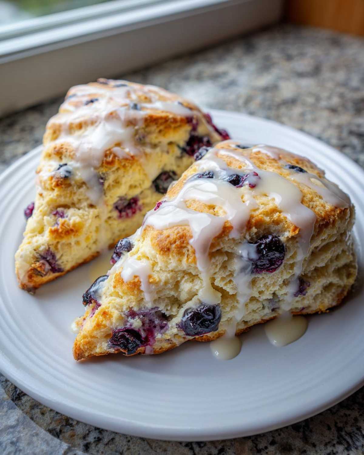 Two freshly baked blueberry scones drizzled with a sweet white glaze, resting on a white plate.