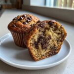 Close-up of a moist banana chocolate chip muffin cut in half showing rich chocolate chips inside.