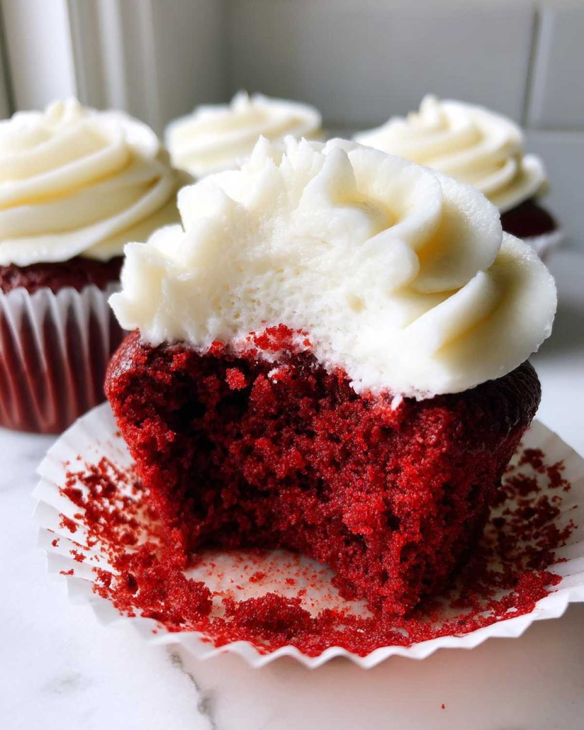 A close-up of a red velvet cupcakes with a bite taken out, showing the moist red crumb and thick cream cheese frosting.