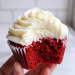 Close-up of a hand holding a red velvet cupcake with a bite taken out, showing the moist red cake and cream cheese frosting.