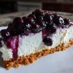 A close-up of a thick slice of creamy blueberry cheesecake with a graham cracker crust and rich blueberry topping.