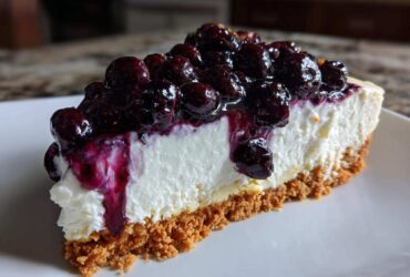 A close-up of a thick slice of creamy blueberry cheesecake with a graham cracker crust and rich blueberry topping.