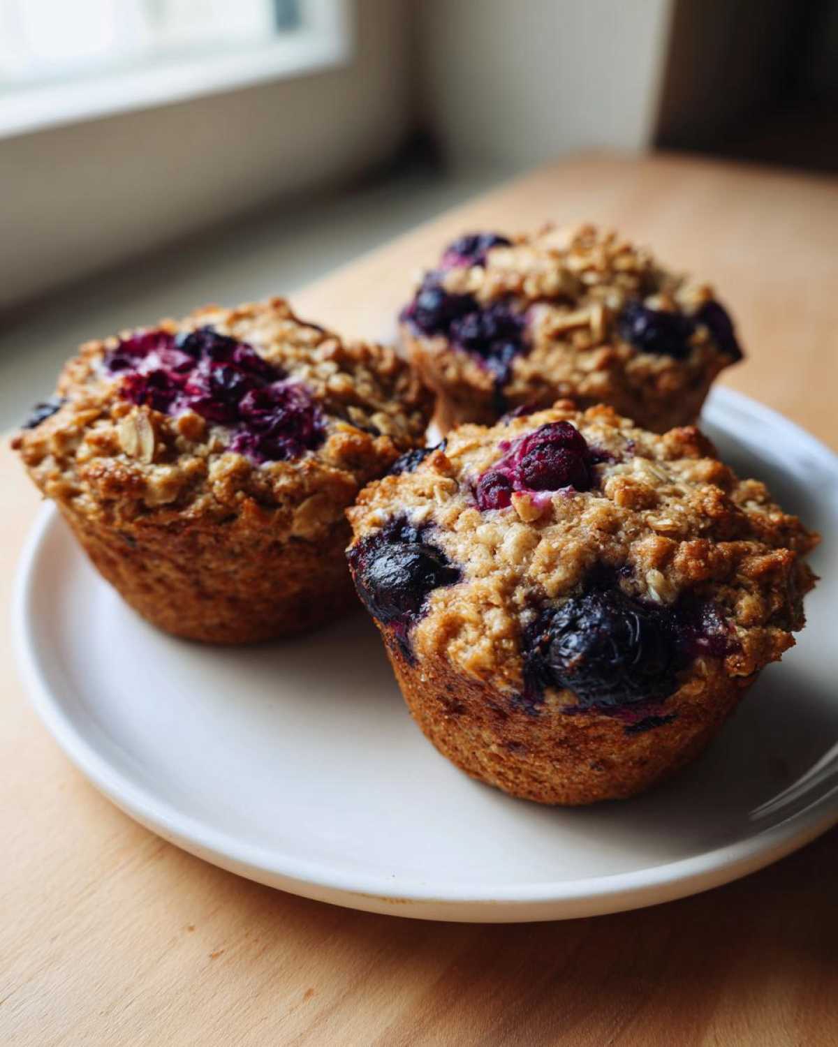 Close-up of three golden brown blueberry oatmeal muffins with crumb topping served on a white plate.
