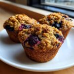 Three freshly baked blueberry oatmeal muffins with a crunchy, golden crumb topping sitting on a white plate.