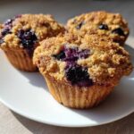 Three golden blueberry oatmeal muffins with a crunchy streusel topping sitting on a white plate.