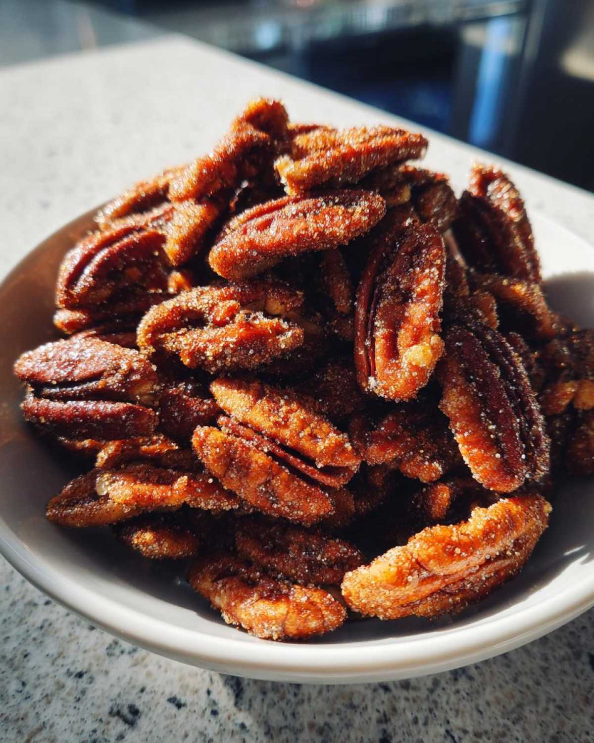 Close-up of a white bowl filled with glistening, caramelized cinnamon sugar pecans, sparkling with sugar crystals.