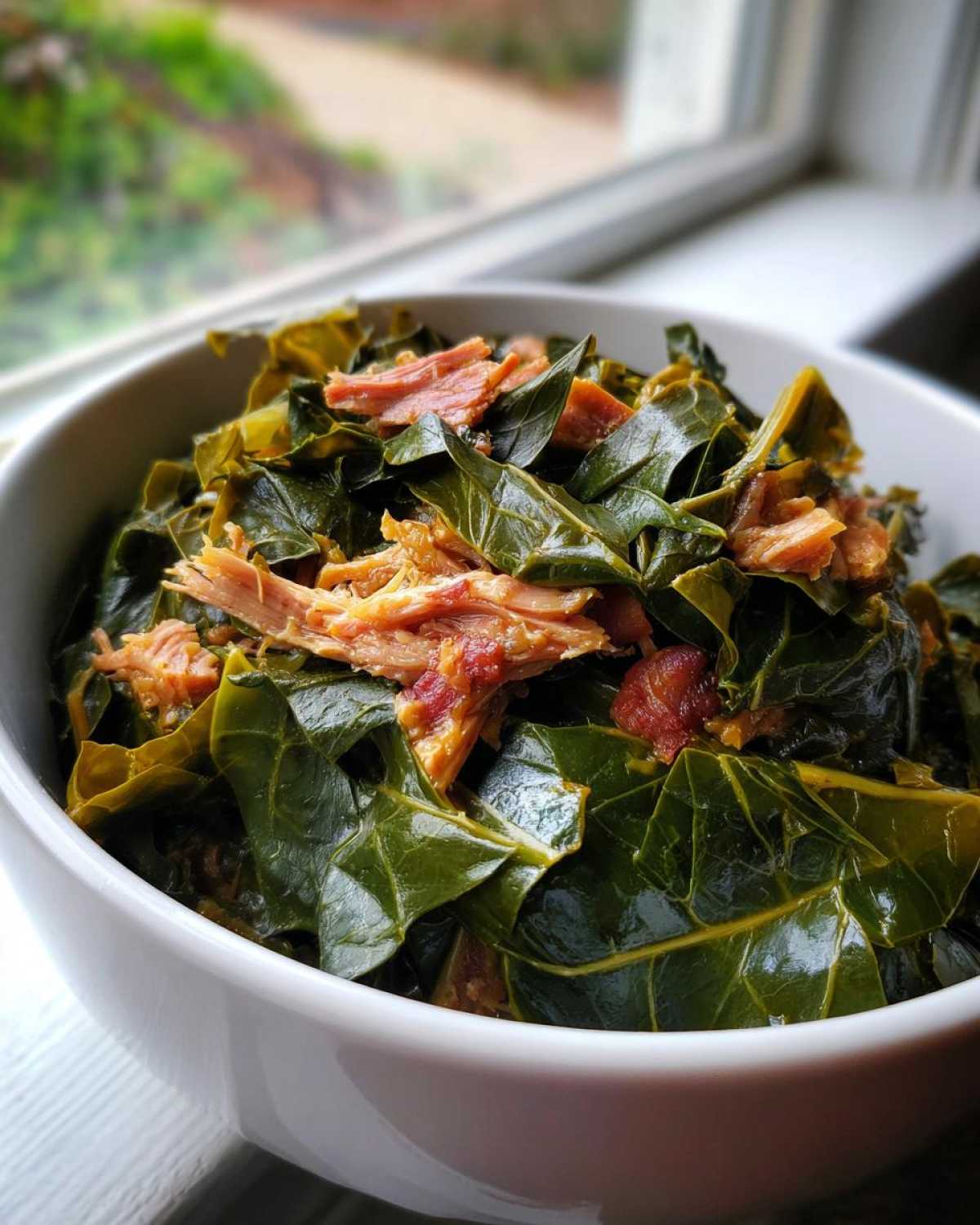 Close-up of a white bowl filled with tender, dark green collard greens mixed with shredded smoked pork.