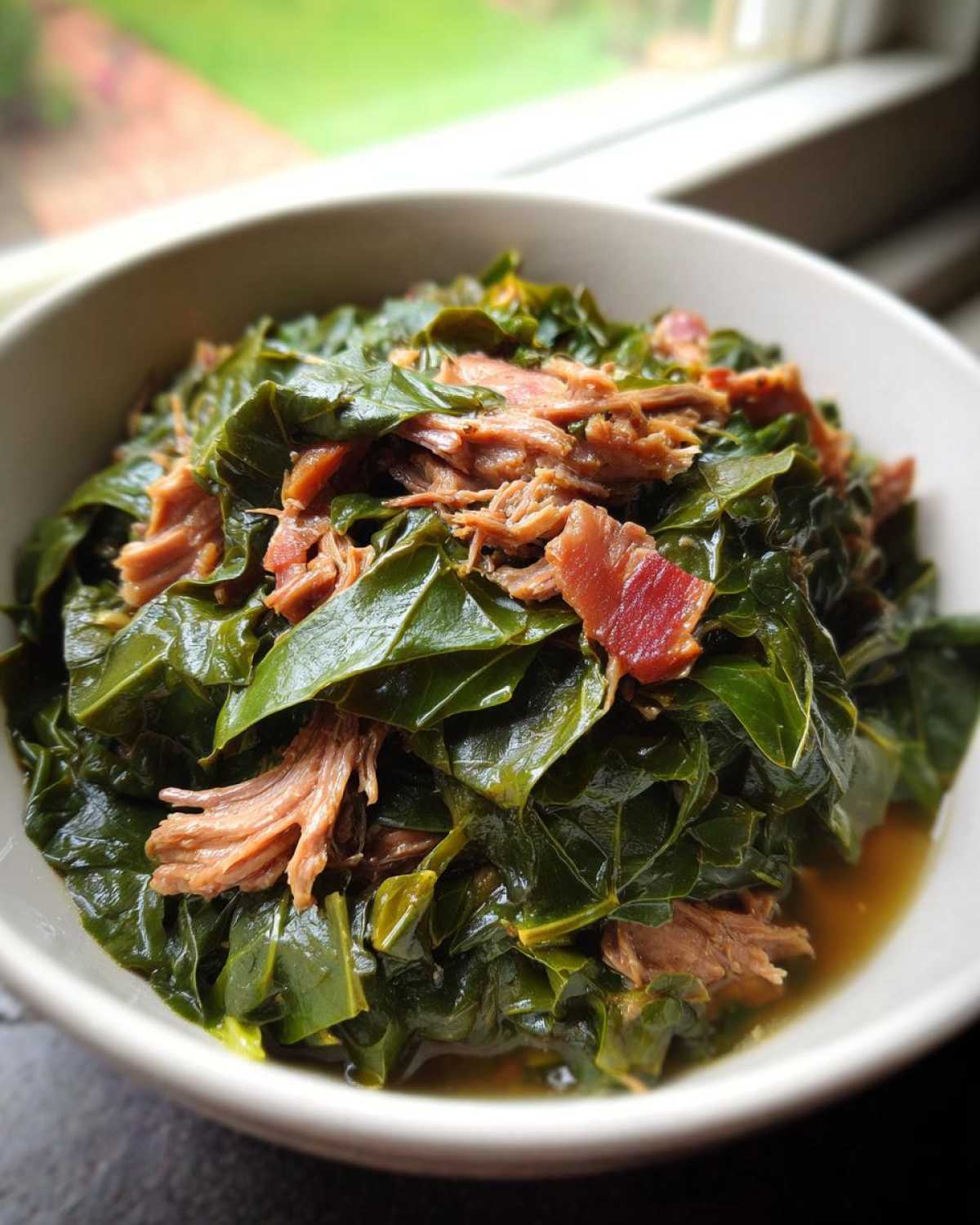 A close-up of tender, dark green collard greens served in a white bowl, topped with shredded smoked ham or bacon.